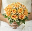In this elegant, hand-gathered bridal posy the apricot roses are the unmistakable protagonists: warm, creamy petals unfolding in tight spirals of peach and soft gold. Between and around each rose, clusters of tiny white gypsophila float like spun sugar, their fine, cloud-like texture providing a breathy contrast that highlights every curve and shadow of the petals. Rich, glossy green foliage wraps the base and edges of the design, shaping the silhouette and lending a fresh, botanical depth to the composition. The light in the photograph falls tenderly across the blooms, picking out warm highlights on the outer petals and casting the inner whorls into a softer, honeyed glow, which makes the whole bouquet feel illuminated from within. To the hand the blossoms would be velvety and cool; the air would carry a delicate, nostalgic perfume-soft tea-rose sweetness tempered by the clean lift of gypsophila and cut foliage. This is the sort of bridal bouquet we prepare for small, thoughtful weddings across Brent Cross and nearby Cricklewood, a piece that reads as both timeless and intimate. Created with patient skill and an eye for proportion, it communicates reassurance and quiet joy, suitable for a ceremony in a local chapel or a relaxed register office celebration. For those seeking reliable craftsmanship and floral balance in the area, Brent Cross Flowers arranges this bouquet with the same steady attention to colour and scent that defines our work.